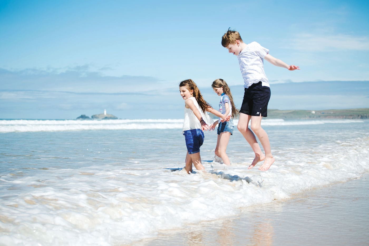 Two girls and a boy on the beach in the shallow white water. The boy is umping, the girls are holding hands. 