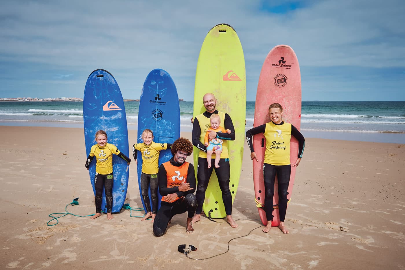 A family at Baleal Surf Camp on their surf holiday. All standing in front of their surfboards. The dad is carrying a baby and the surf instructor is kneeled in front of them