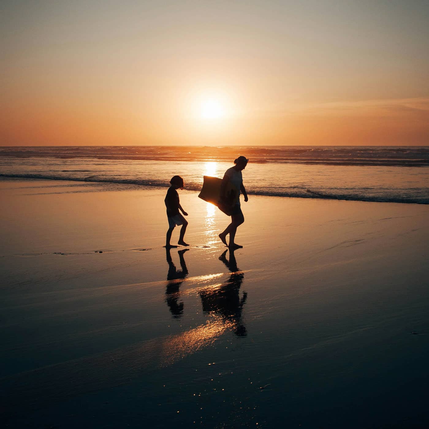 A mum and her son walking along the beach with a bodyboard in the sunset on their family surf holiday