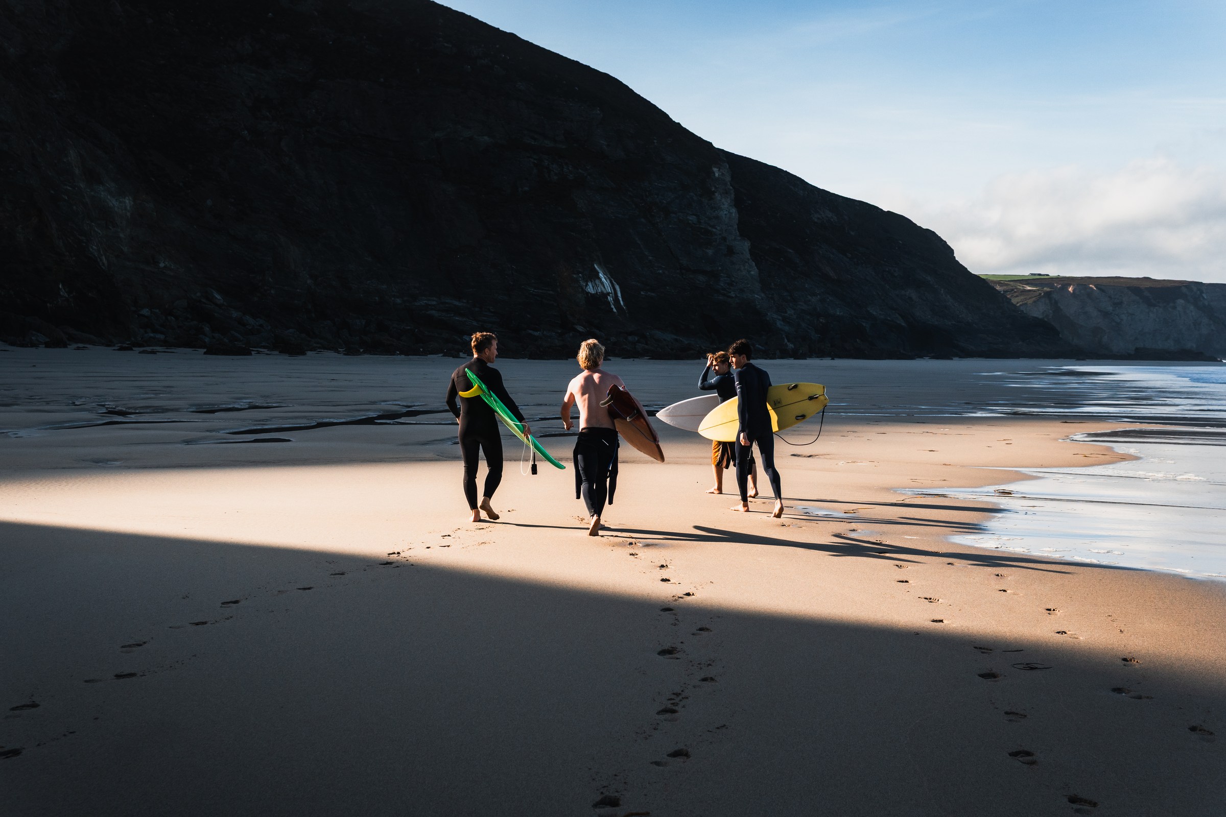 Four surfers carrying boards across a sunlit beach beneath a dark cliff