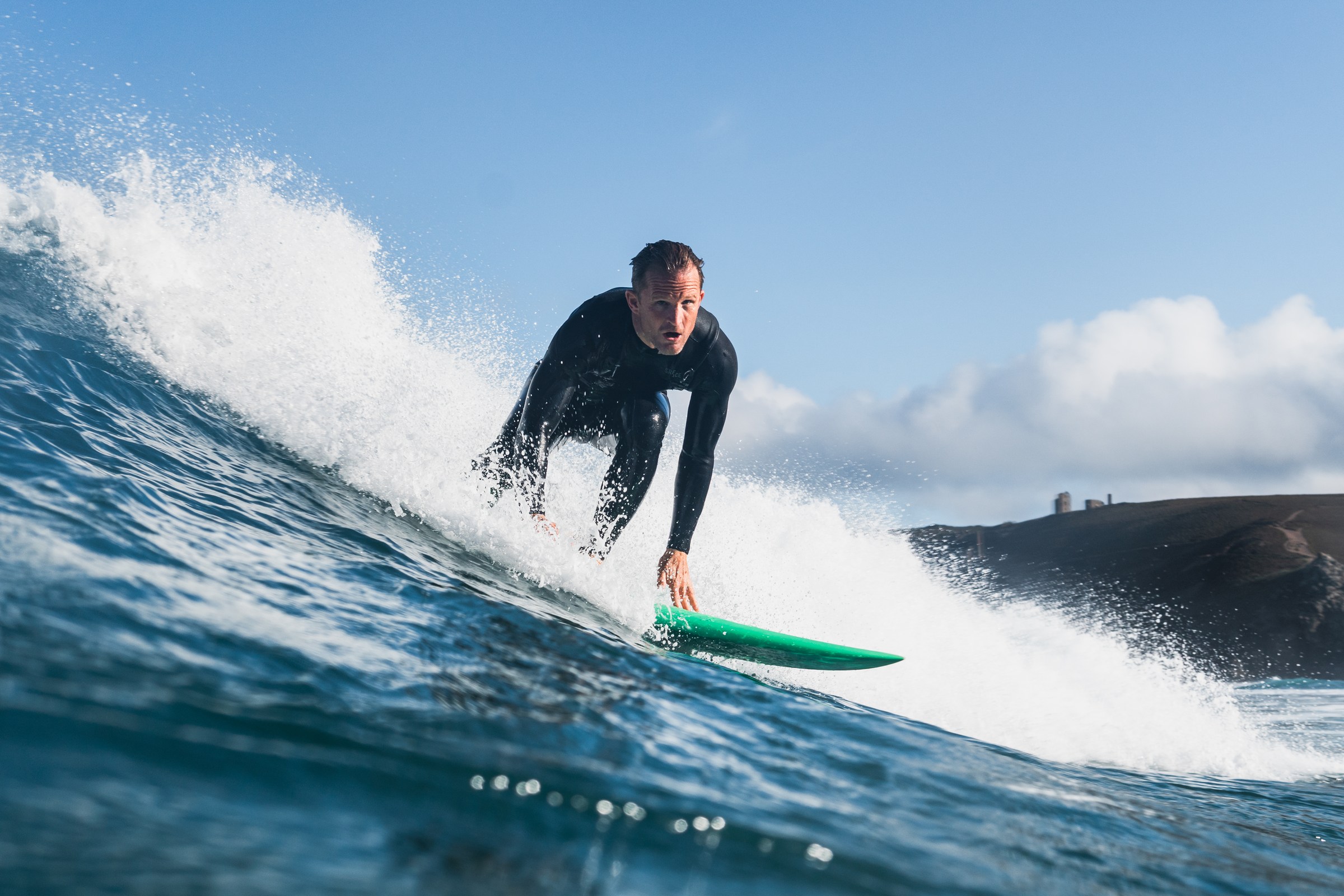 Joel riding a green board down the face of a wave with a stone building on the headland behind