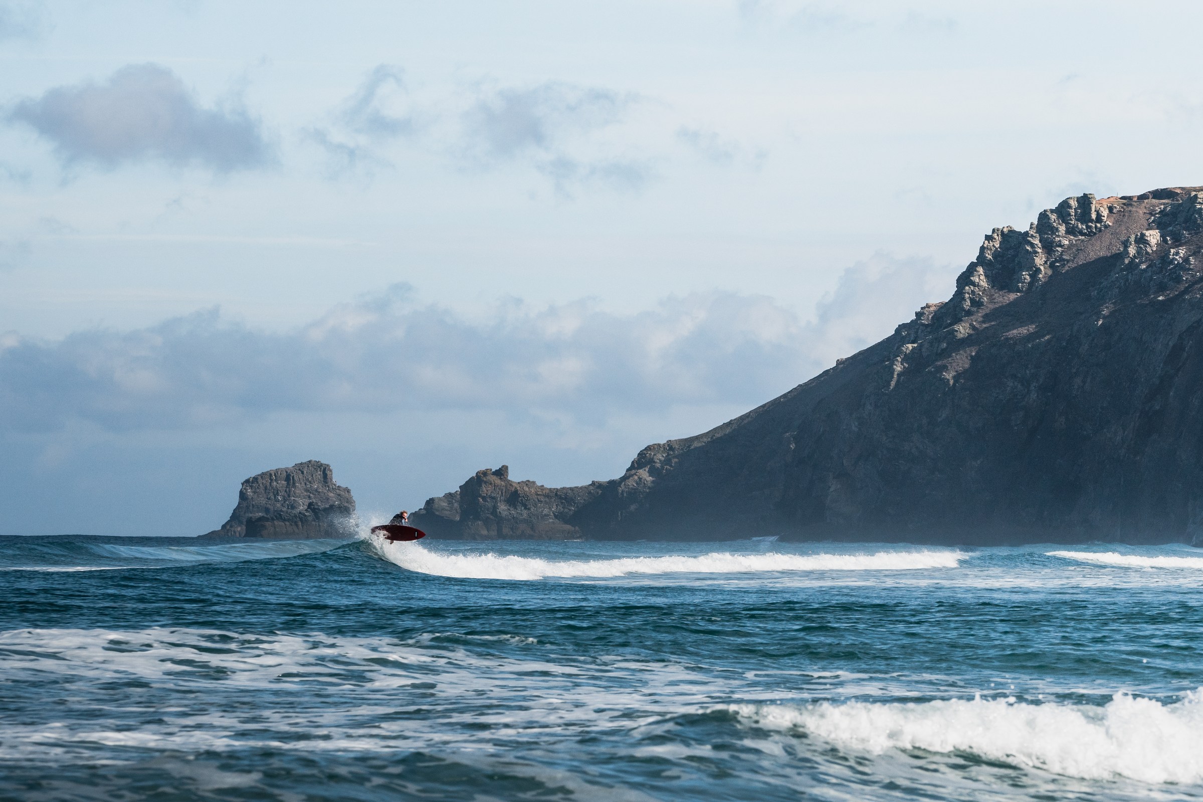 Surfer launching an air off the lip on a red board beneath a rocky headland