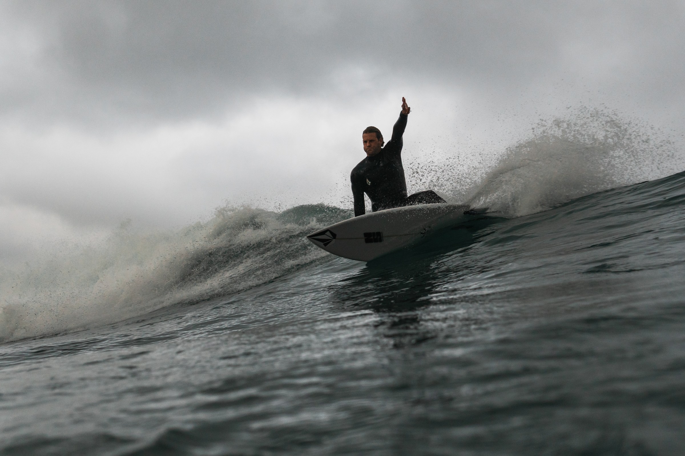 Harry Timson on a white board with one arm raised on a wave under dark cloud