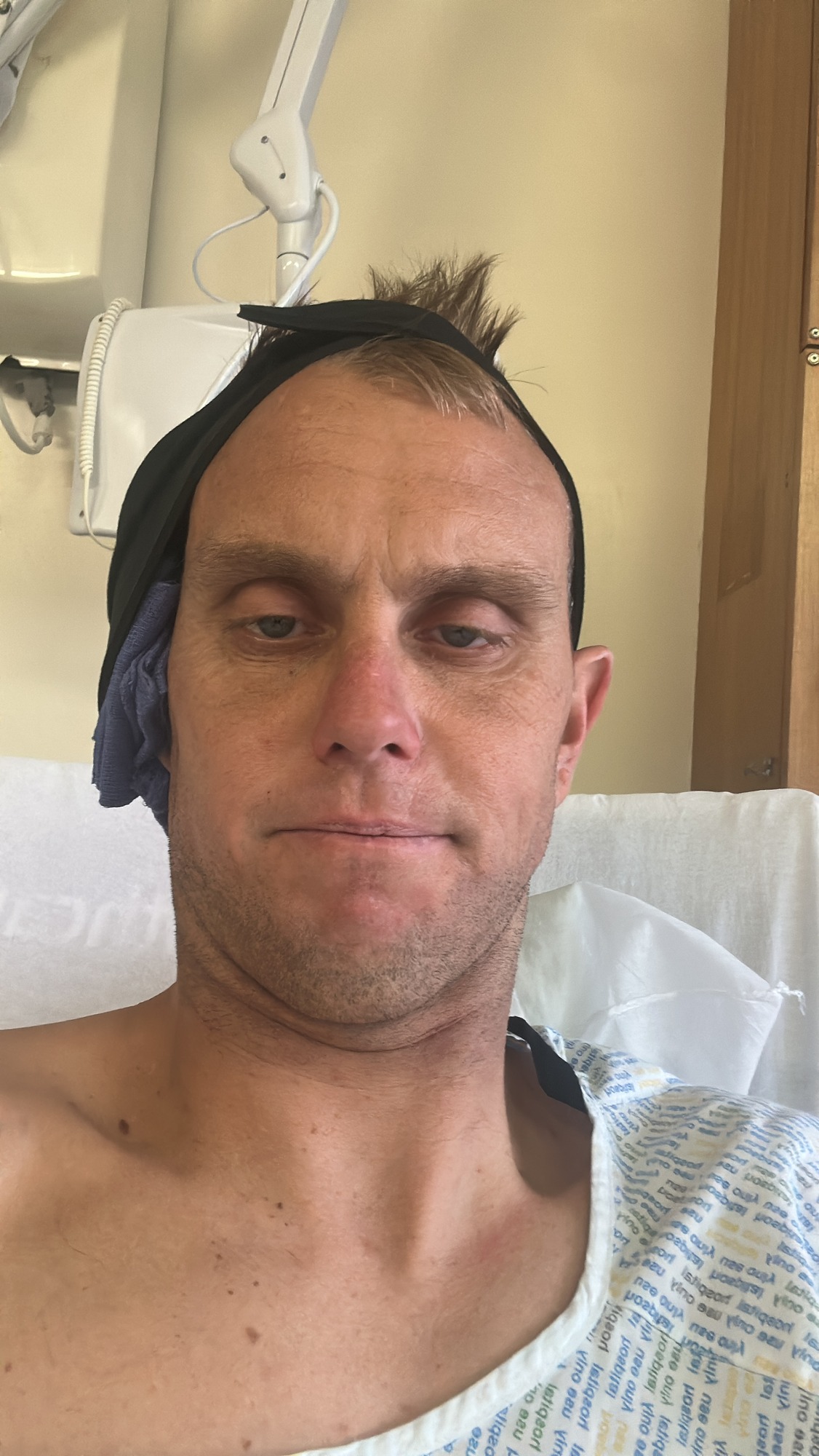 Mark Boyd sitting up in a hospital bed in a gown with a bandage wrapped around his head after ear surgery