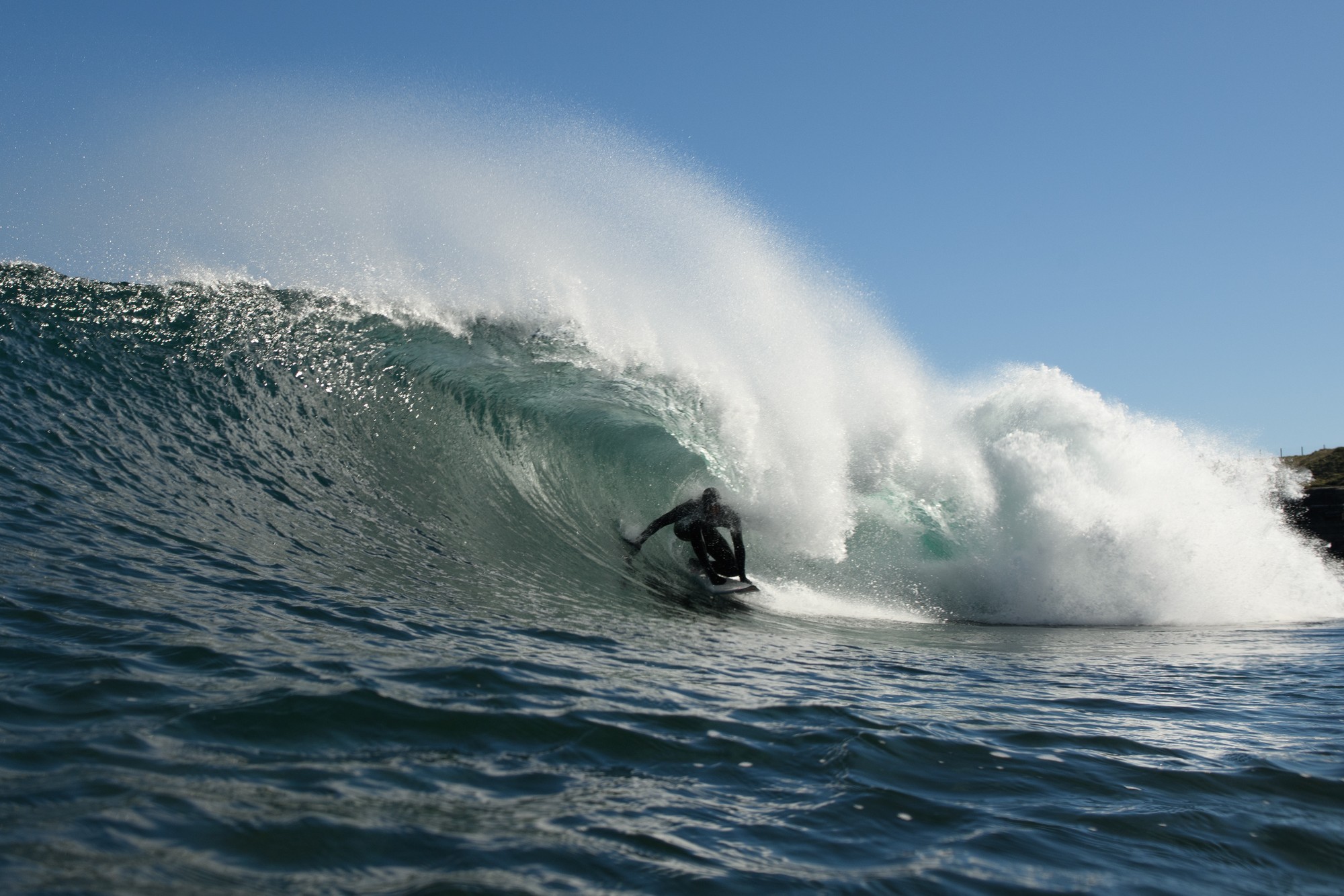 Mark Boyd pulling into a barrel under a plume of spray on a bright blue day