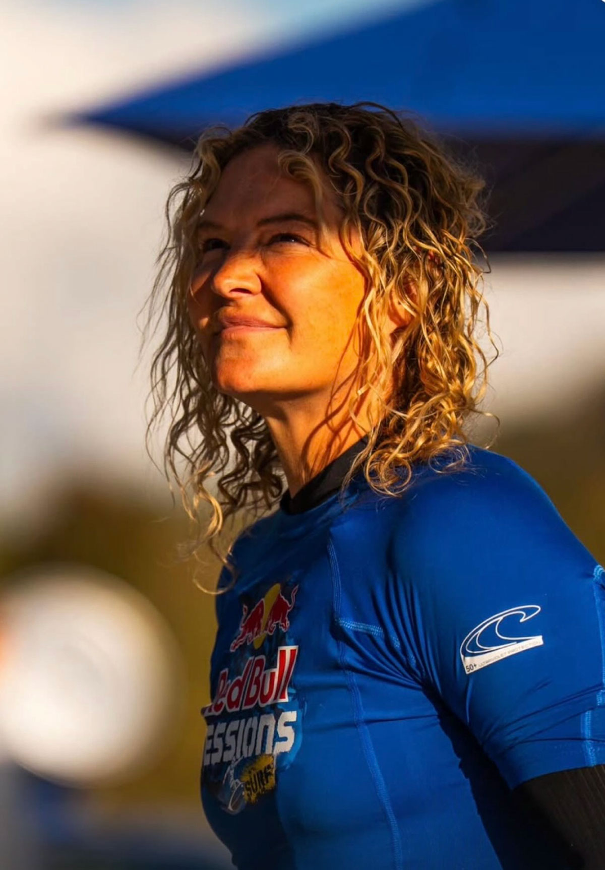 Zoe Smith with wet curly blonde hair, smiling and looking up in low sun, wearing a blue Red Bull Sessions rash vest