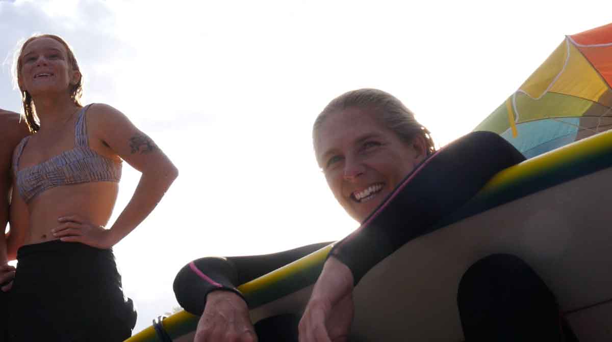 Caity Simmers and Steph Gilmore laughing beside a surfboard, backlit, with a rainbow umbrella behind them