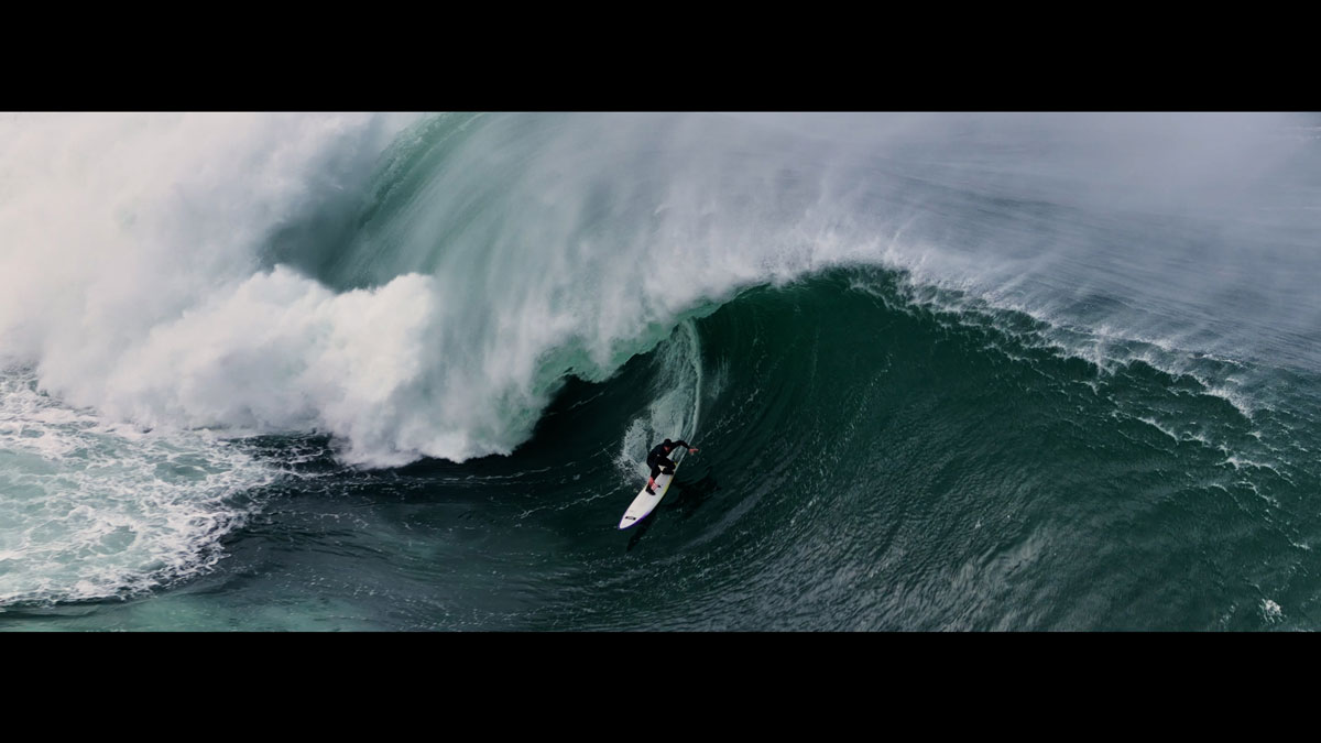 Aerial view of a surfer racing down the face of a huge green wave as the lip throws over behind him