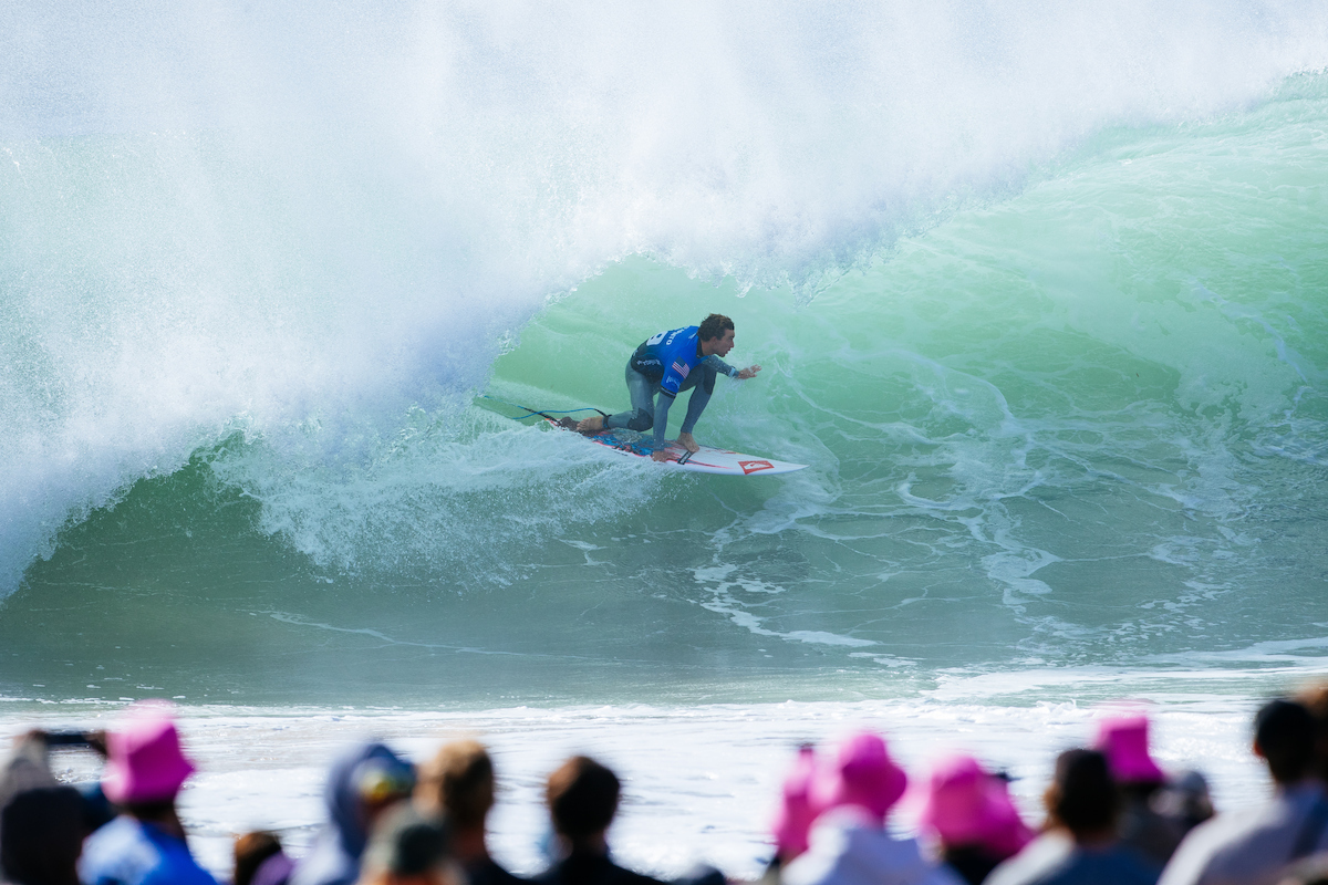Griffin Colapinto in a blue jersey crouched in a heaving green barrel at Supertubos, with spectators in pink hats in the foreground