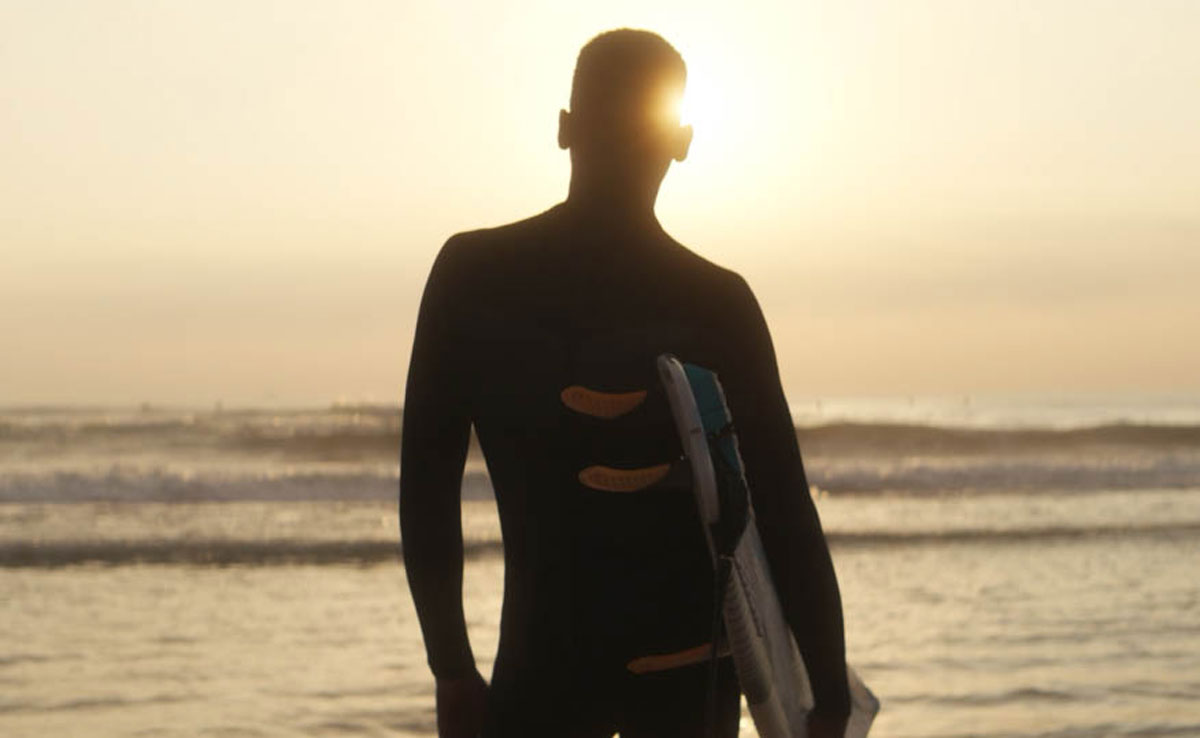 A surfer in a wetsuit, seen from behind in silhouette, holds his board under one arm and faces the sea at sunset