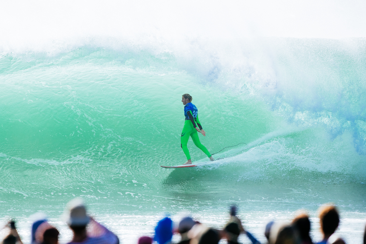 Molly Picklum in a green wetsuit and blue jersey standing tall inside a large turquoise barrel at Supertubos, with spectators in the foreground