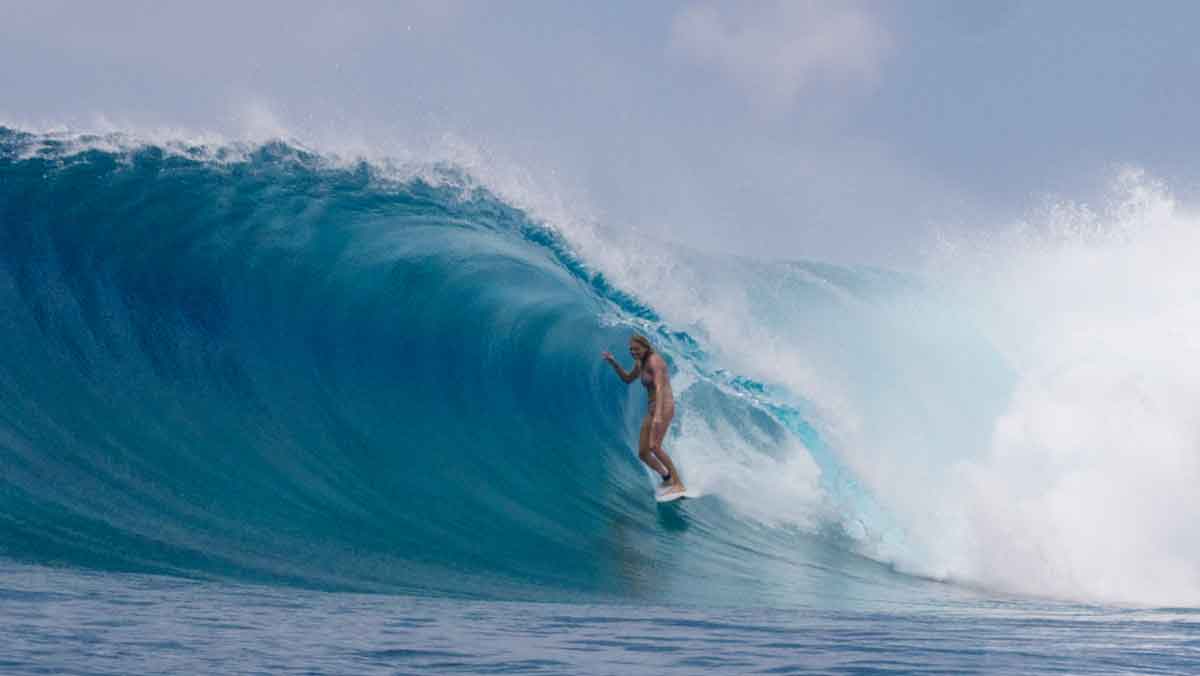 Steph Gilmore standing tall in a large blue barrel, one hand trailing towards the face