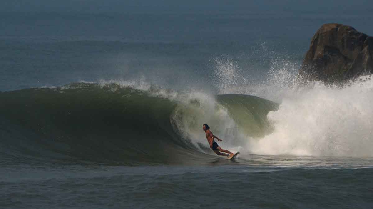 Derrick Disney in boardshorts racing along a green right-hander beside a large rock