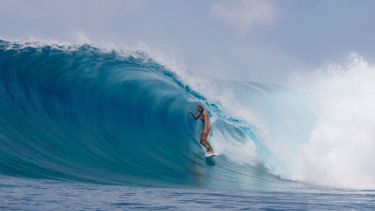 A surfer in a bikini stands tall inside a big blue tropical barrel