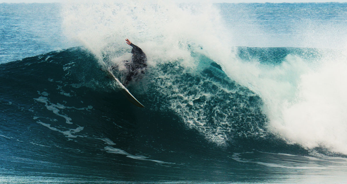 A surfer in a wetsuit drives his board vertically up the lip of a heavy blue wave, spray exploding around him