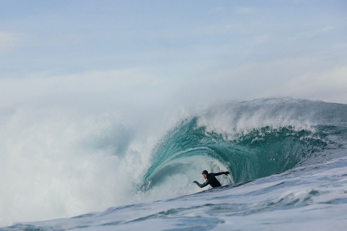 Jayce Robinson in a hooded wetsuit and gloves sets his arms inside a thick green cold-water barrel under a pale sky