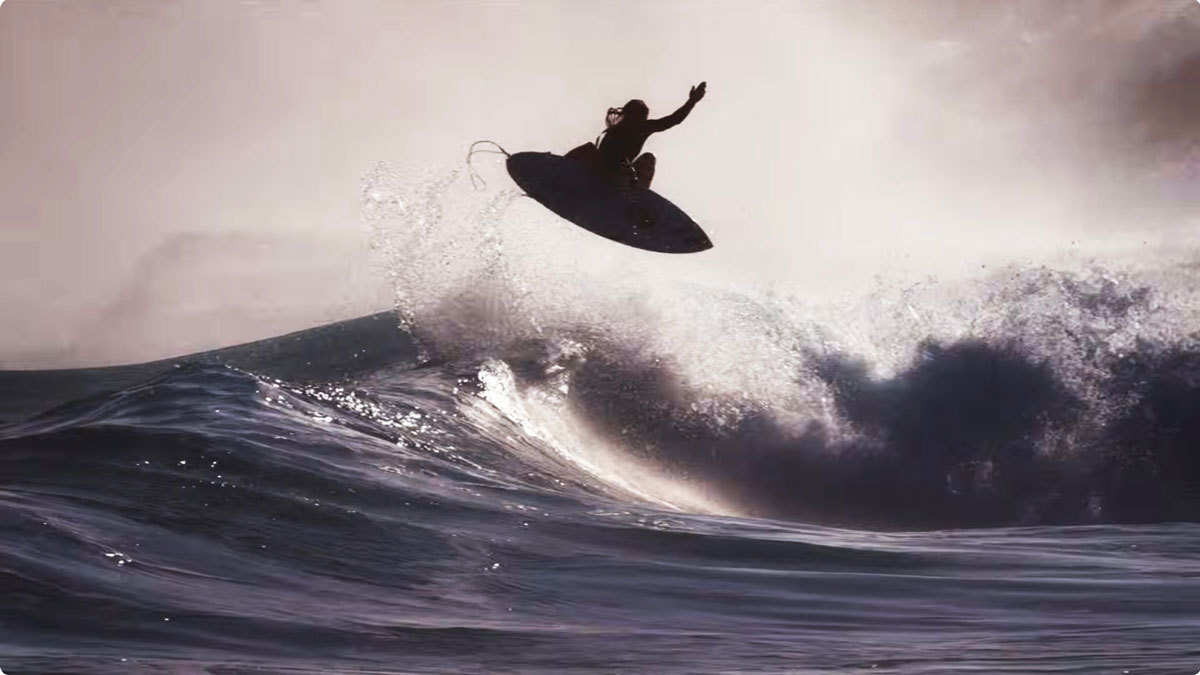 A surfer in silhouette flies through the air above a dark breaking wave, spray lit from behind