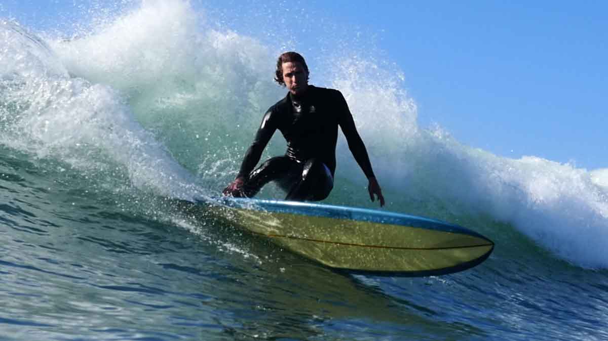 Bryce Young in a black wetsuit crouched low on a yellow and blue longboard, spray rising off the lip behind him