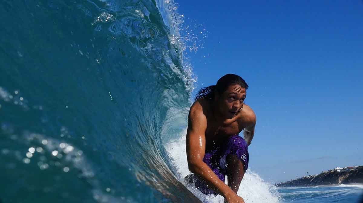 Kobe in purple boardshorts crouched in a clear blue barrel, shot from inside the wave, with a cliff in the distance