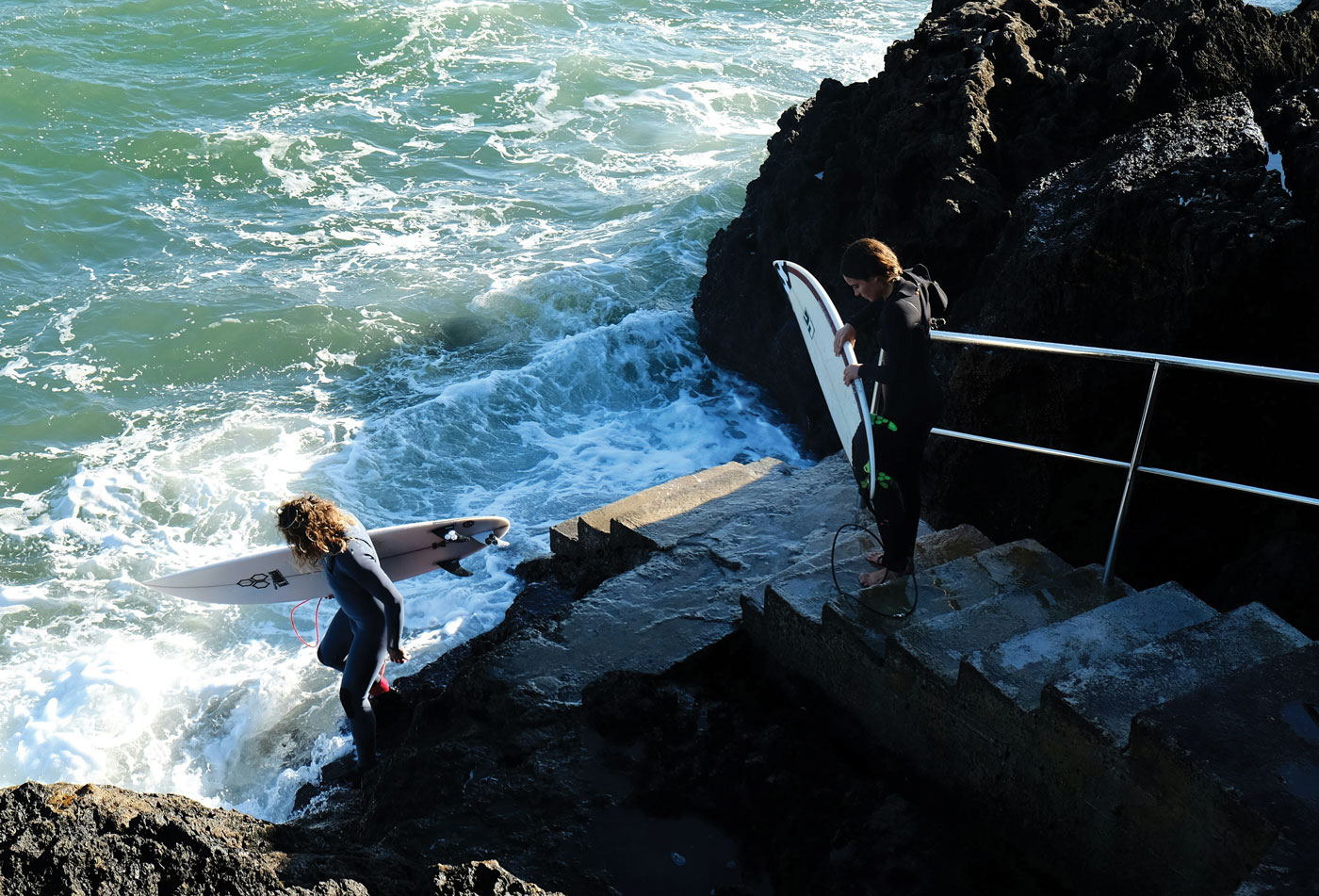 Two surfers in wetsuits carry their boards down stone steps to a churning sea