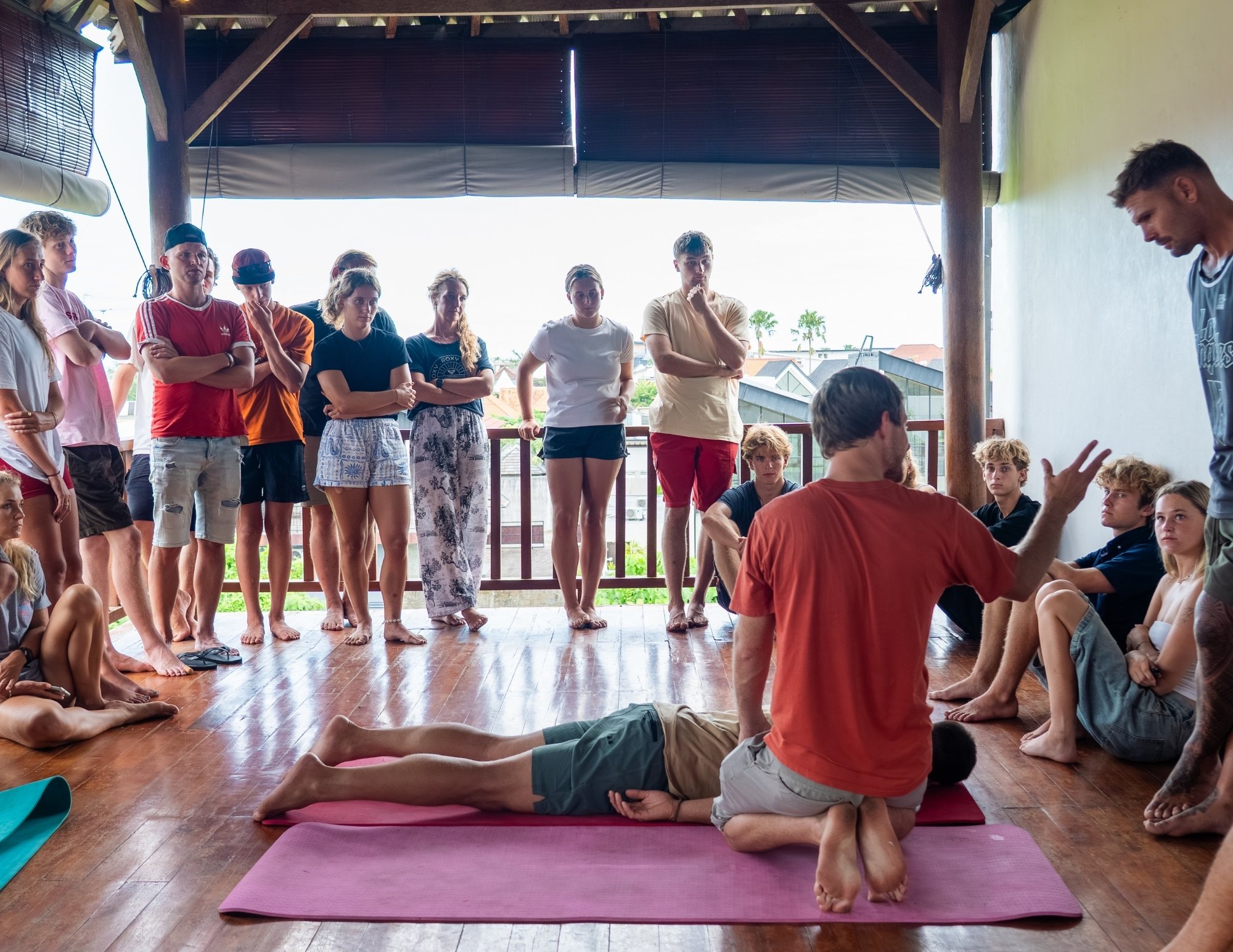 An instructor demonstrates a technique on a person lying on a mat while a group watches on a wooden terrace