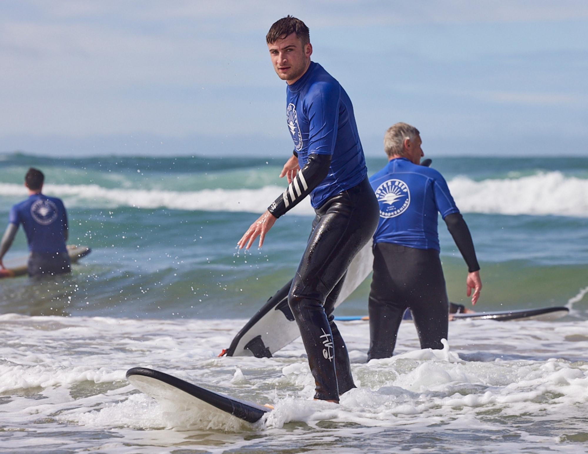 A surfer in a blue Outer Reef rash vest and black wetsuit stands on a board in the white water, looking back towards the camera