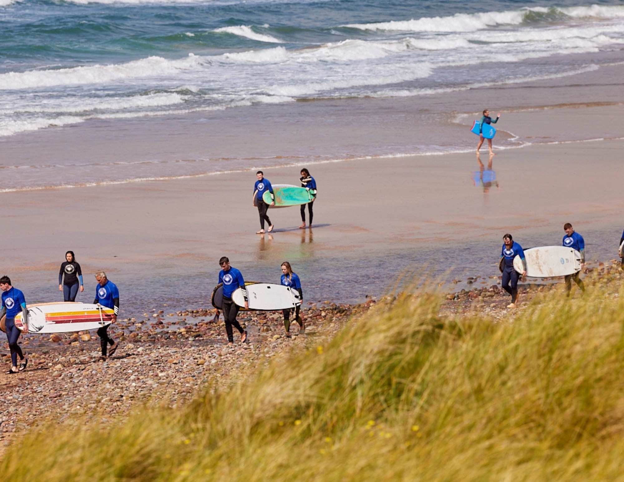 Surfers in blue Outer Reef rash vests carry boards along a pebbly beach beside dune grass