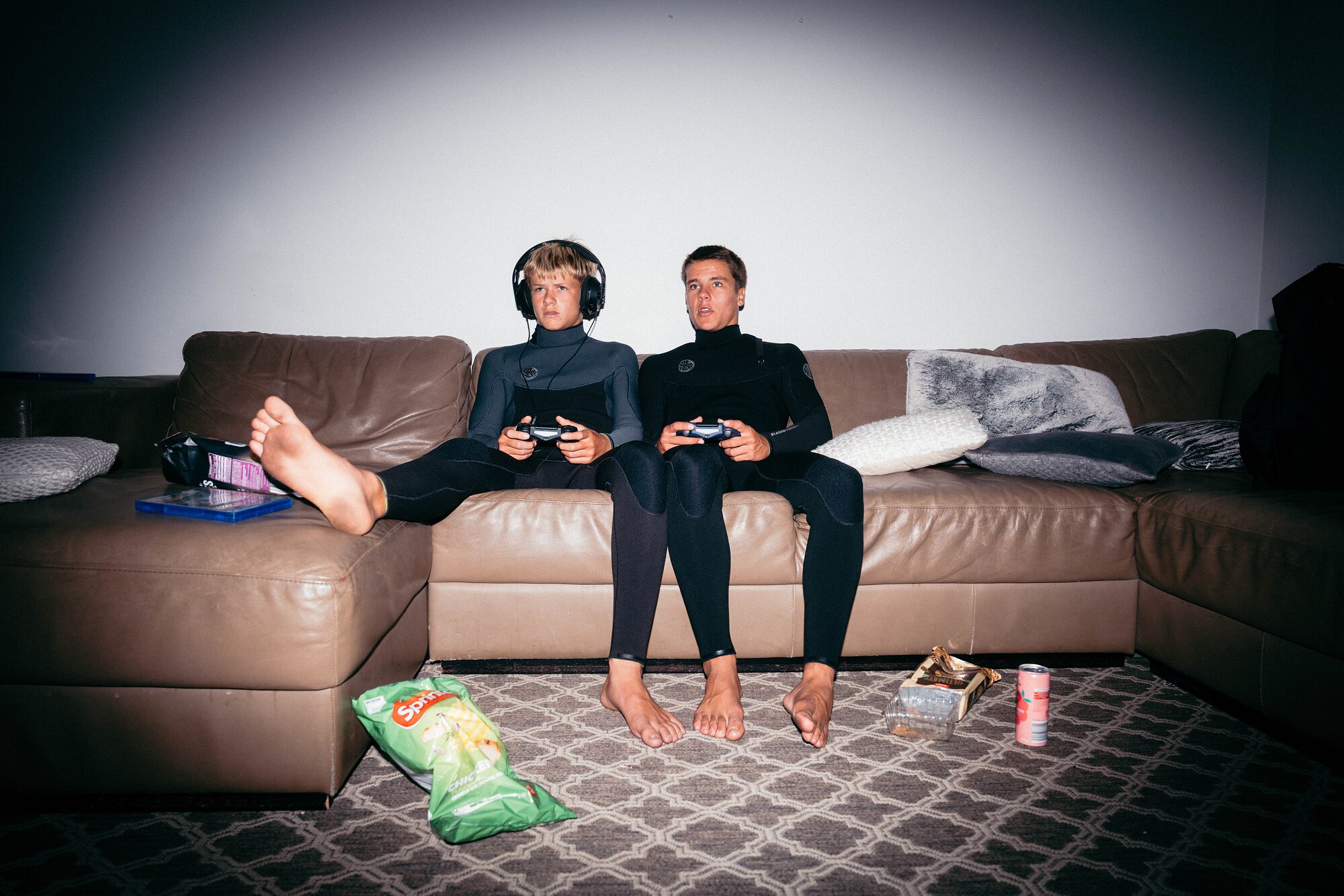 Two young surfers in wetsuits playing video games on a leather sofa