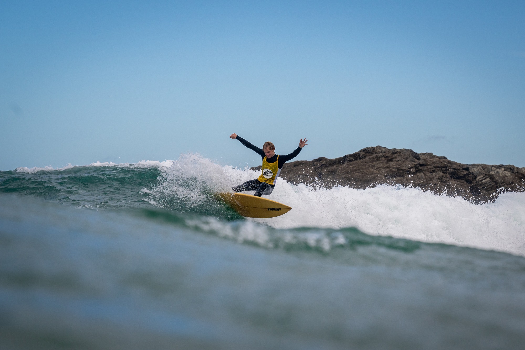 Surfer in a yellow bib with both arms out on a small wave in front of a rock