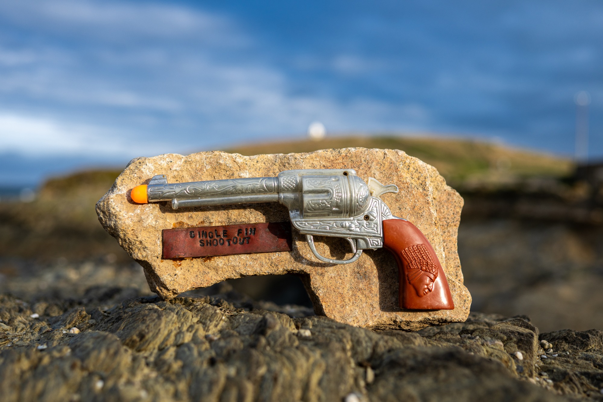 Toy revolver mounted on a stone with a Single Fin Shootout plaque, set against the coast