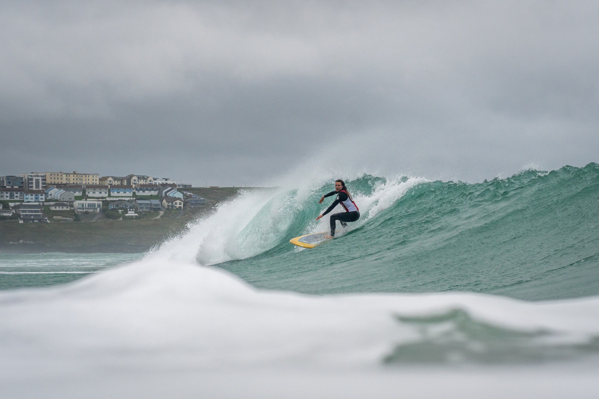 Surfer drawing a line along a green wave beneath a grey sky with Newquay buildings behind