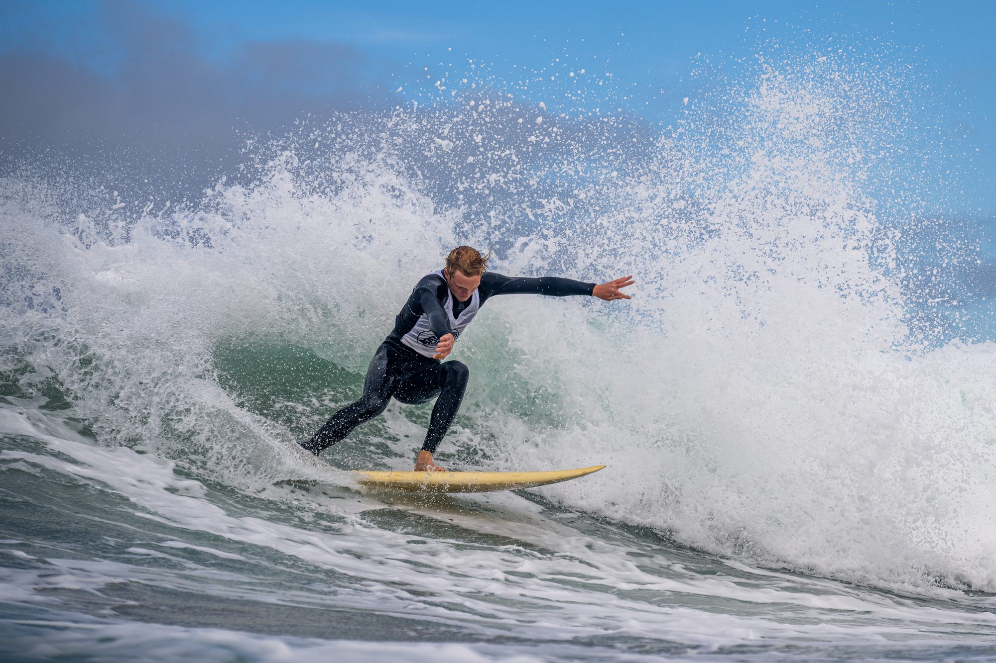 Surfer in a black wetsuit and white bib carving through a burst of spray on a yellow single fin